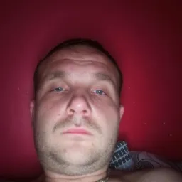 Mężczyzna z bliska, selfie na tle czerwonej ściany, widoczny srebrny łańcuszek na szyi.