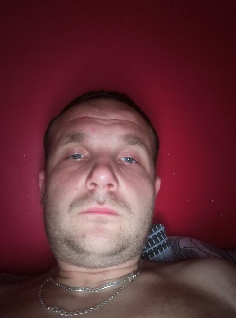 Mężczyzna z bliska, selfie na tle czerwonej ściany, widoczny srebrny łańcuszek na szyi.