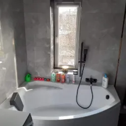 Nowoczesna łazienka z wanną narożną i armaturą w kolorze czarnym, wykończona szarymi płytkami imitującymi marmur, z oknem na tle drzew bez liści i kolorowymi butelkami kosmetyków na wannie.