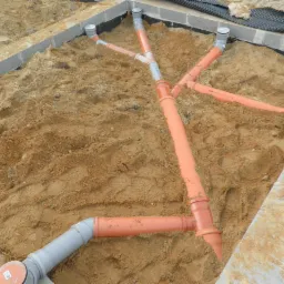 Pomarańczowe i szare rury kanalizacyjne ułożone w ziemi, przygotowane do podłączenia przydomowej oczyszczalni ściek&oacute;w, widoczne betonowe obrzeża.