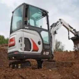 Kompaktowa minikoparka Bobcat E19 w trakcie pracy na placu budowy, z łyżką pełną ziemi, widoczna w tle zieleń.