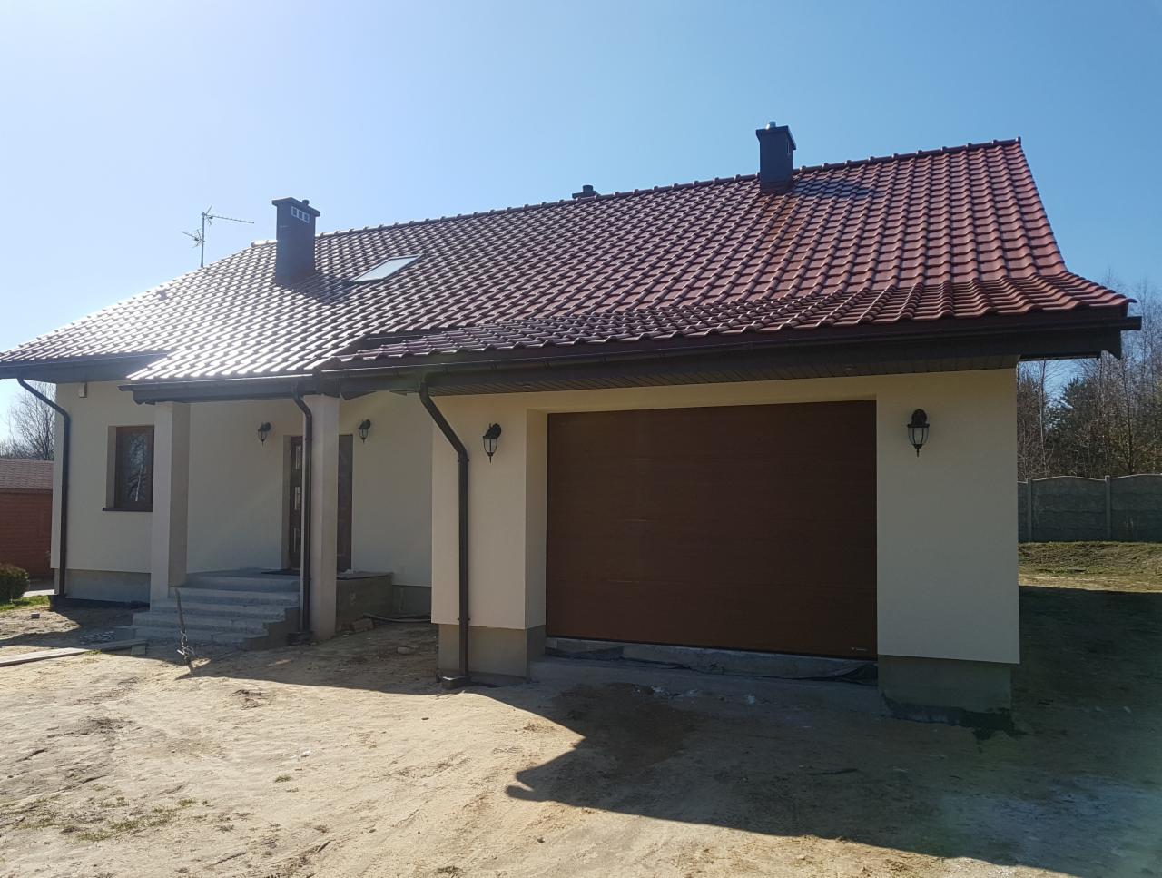 Jednopiętrowy dom z brązową dachówką, beżową elewacją i brązową bramą garażową, oświetlony zewnętrznymi lampami, z widocznymi schodami wejściowymi i częściowo zagospodarowanym terenem.