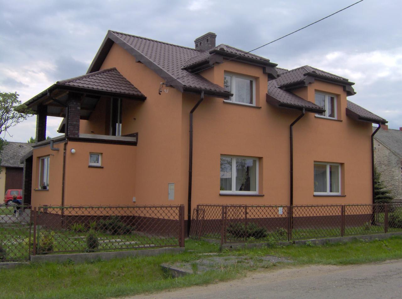 Dwukondygnacyjny dom z balkonem i brązowym dachem, pomalowany na kolor brzoskwiniowy, otoczony niskim, metalowym płotem.