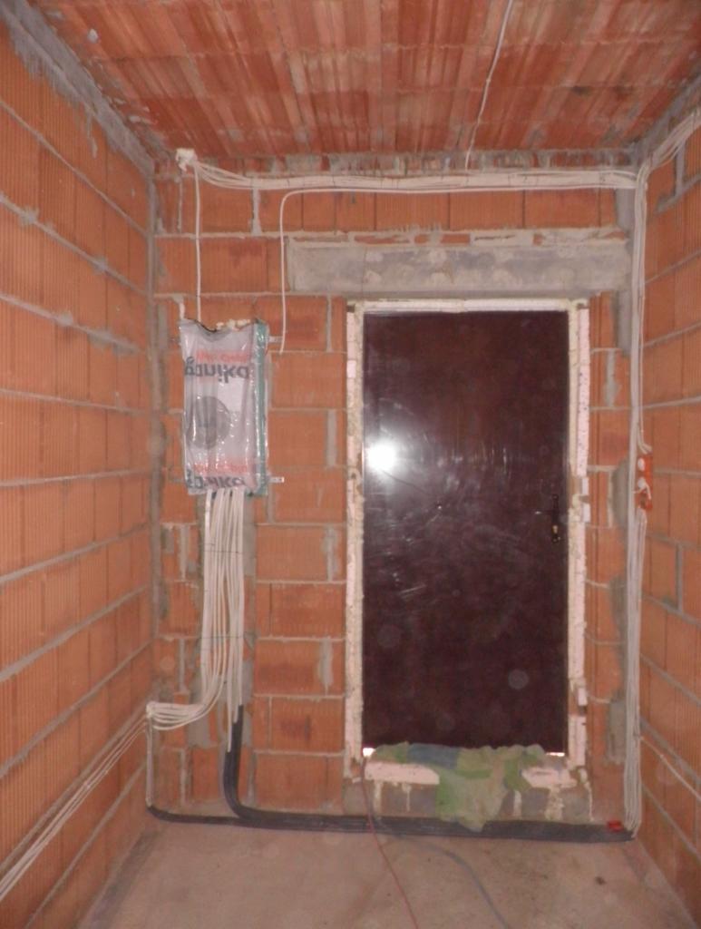 Surowy stan wnętrza: ściany z czerwonej cegły dziurawki, wystające przewody elektryczne i puszka instalacyjna, ciemne drzwi wewnętrzne, betonowa podłoga.