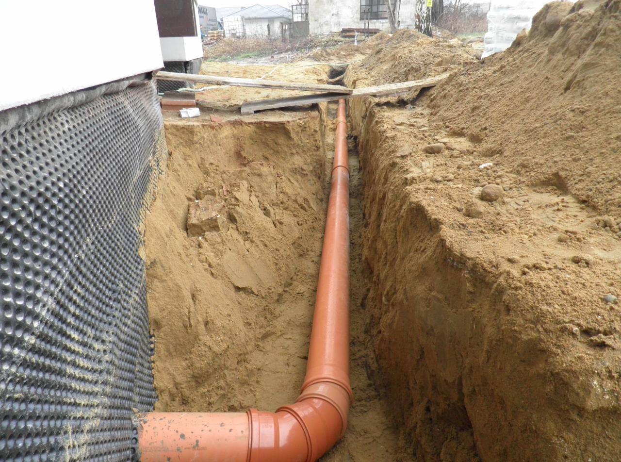 Pomarańczowa rura kanalizacyjna ułożona w wykopie ziemnym wzdłuż fundamentu budynku, zabezpieczonego folią kubełkową.