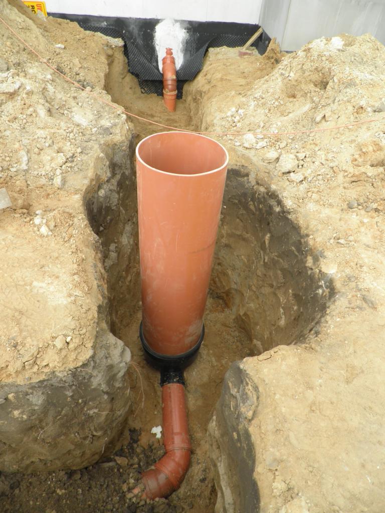 Pomarańczowa rura kanalizacyjna w wykopie ziemnym, z widocznym kolankiem i połączeniem z drenażem, zabezpieczona membraną hydroizolacyjną przy białej ścianie fundamentowej.