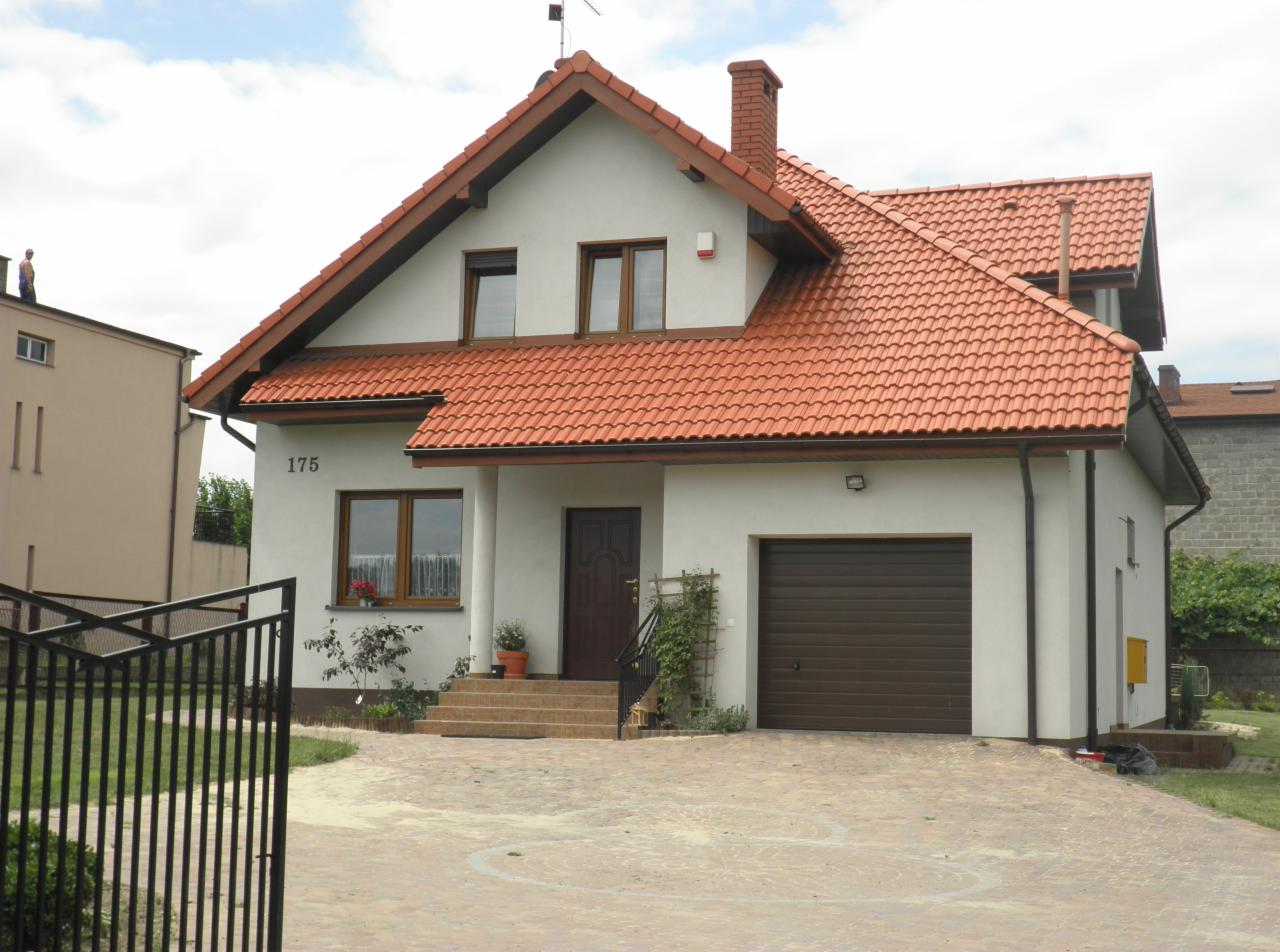 Dwukondygnacyjny dom jednorodzinny z czerwoną dachówką, brązową bramą garażową i numerem 175 na elewacji, widoczny zza czarnego metalowego ogrodzenia.