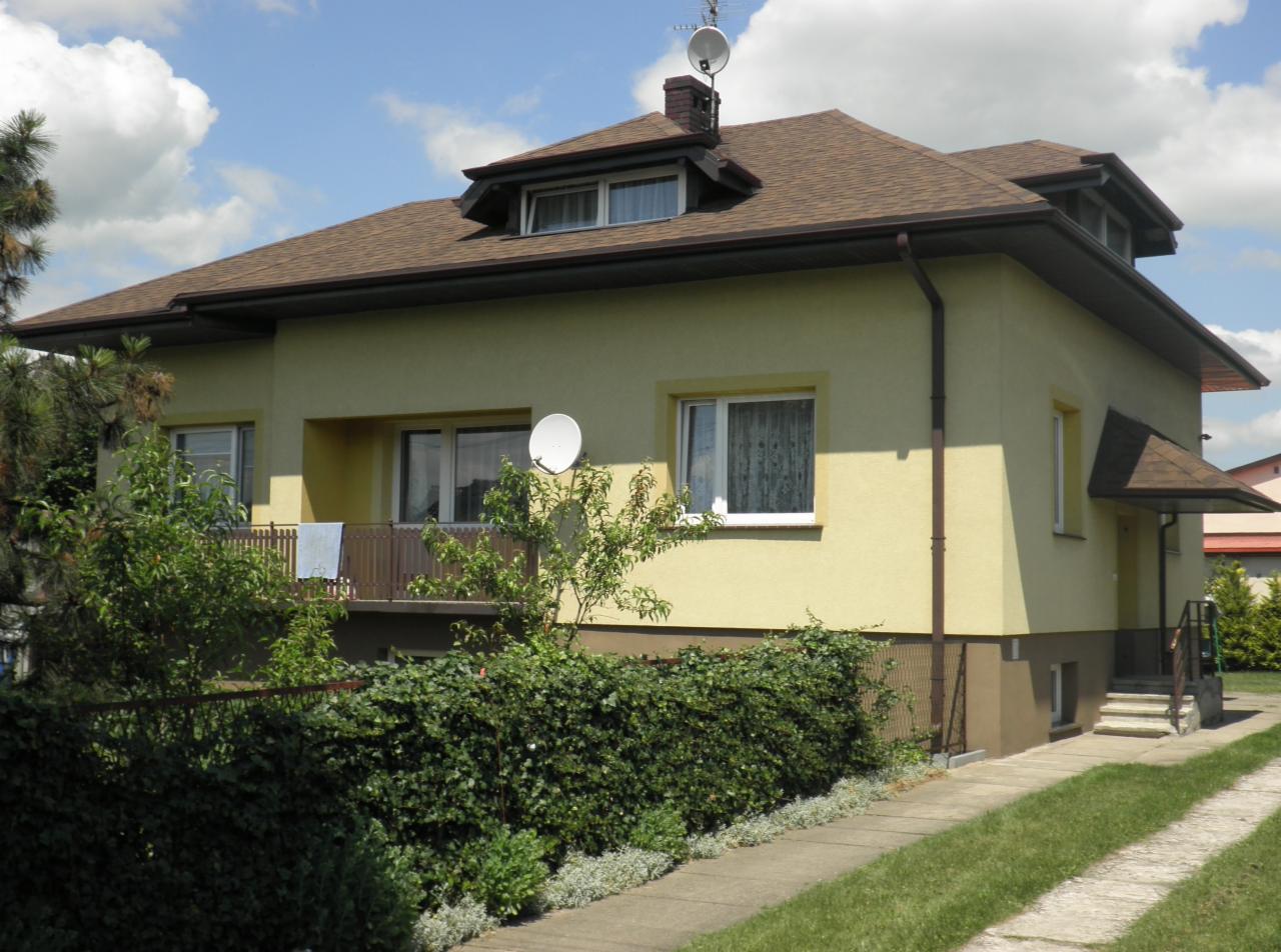 Dwukondygnacyjny dom z brązowym dachem i żółtą elewacją, widoczny balkon z suszącym się ręcznikiem i antena satelitarna.