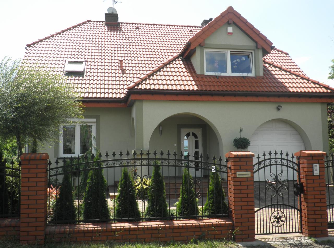 Dwupiętrowy dom z czerwonym dachem i jasną elewacją, widoczny zza ozdobnego, czarnego ogrodzenia z ceglanymi słupkami.