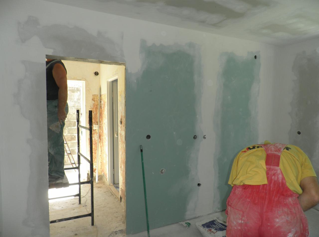 Wnętrze w trakcie szpachlowania płyt gipsowo-kartonowych; widoczny pracownik w czerwonym kombinezonie, drabina oraz ślady szpachli na ścianach i suficie.