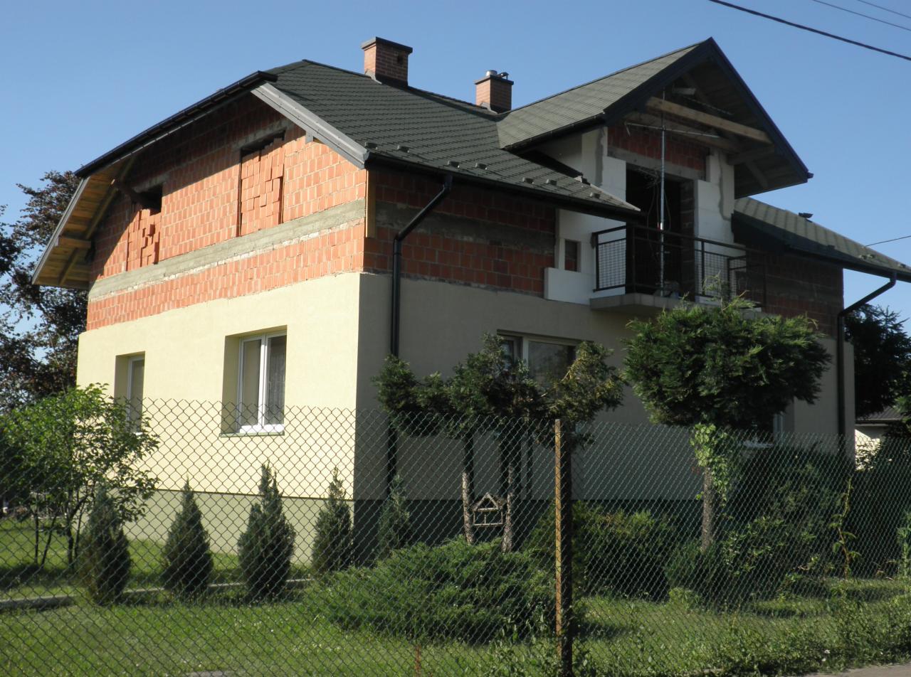 Dwupiętrowy dom w trakcie budowy, z częściowo otynkowaną elewacją, widocznymi cegłami i balkonem z metalową barierką, ogrodzony siatką.