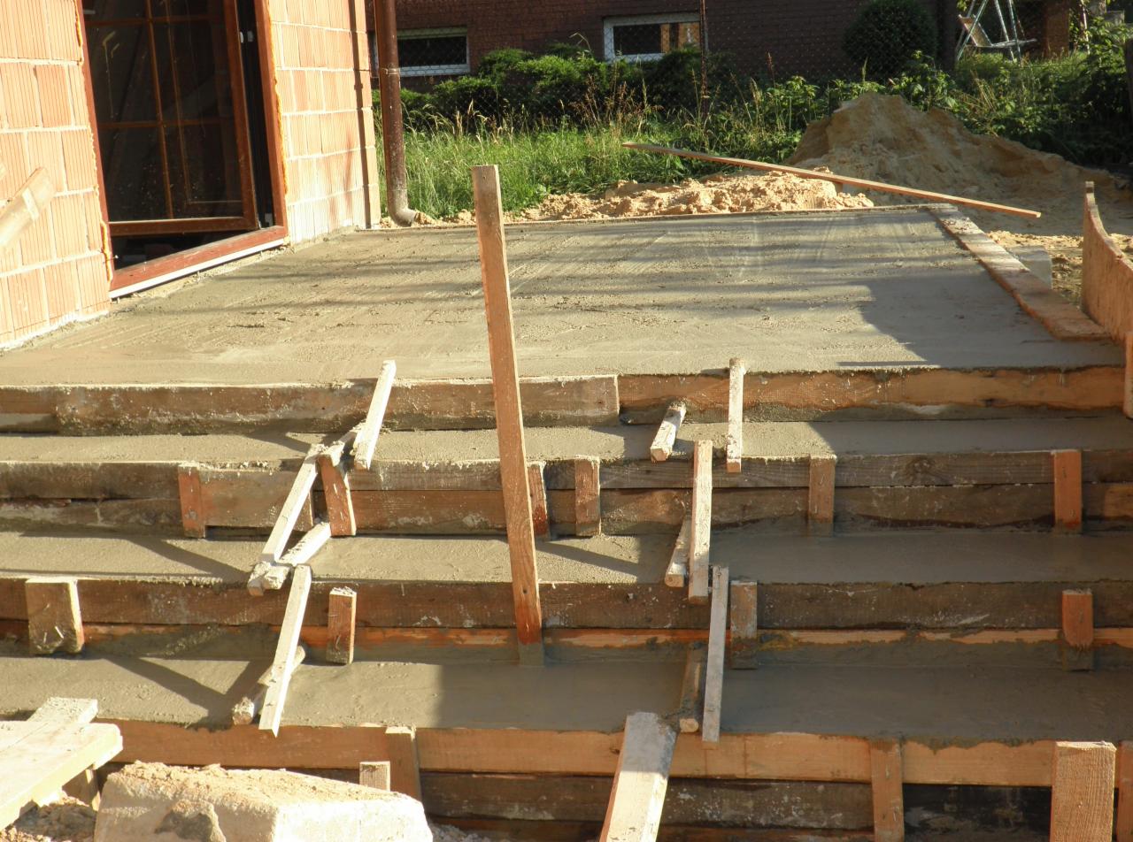 Betonowe schody w trakcie budowy, drewniane szalunki, widoczny fragment ściany budynku z cegły.