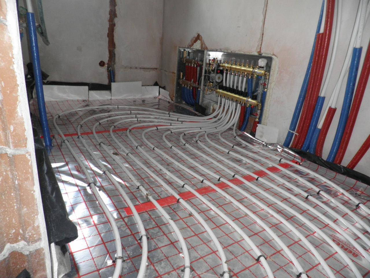 Rozłożone rury ogrzewania podłogowego na folii aluminiowej z kratką, widoczny rozdzielacz z rurami w kolorze czerwonym i niebieskim, surowe ściany w tle.