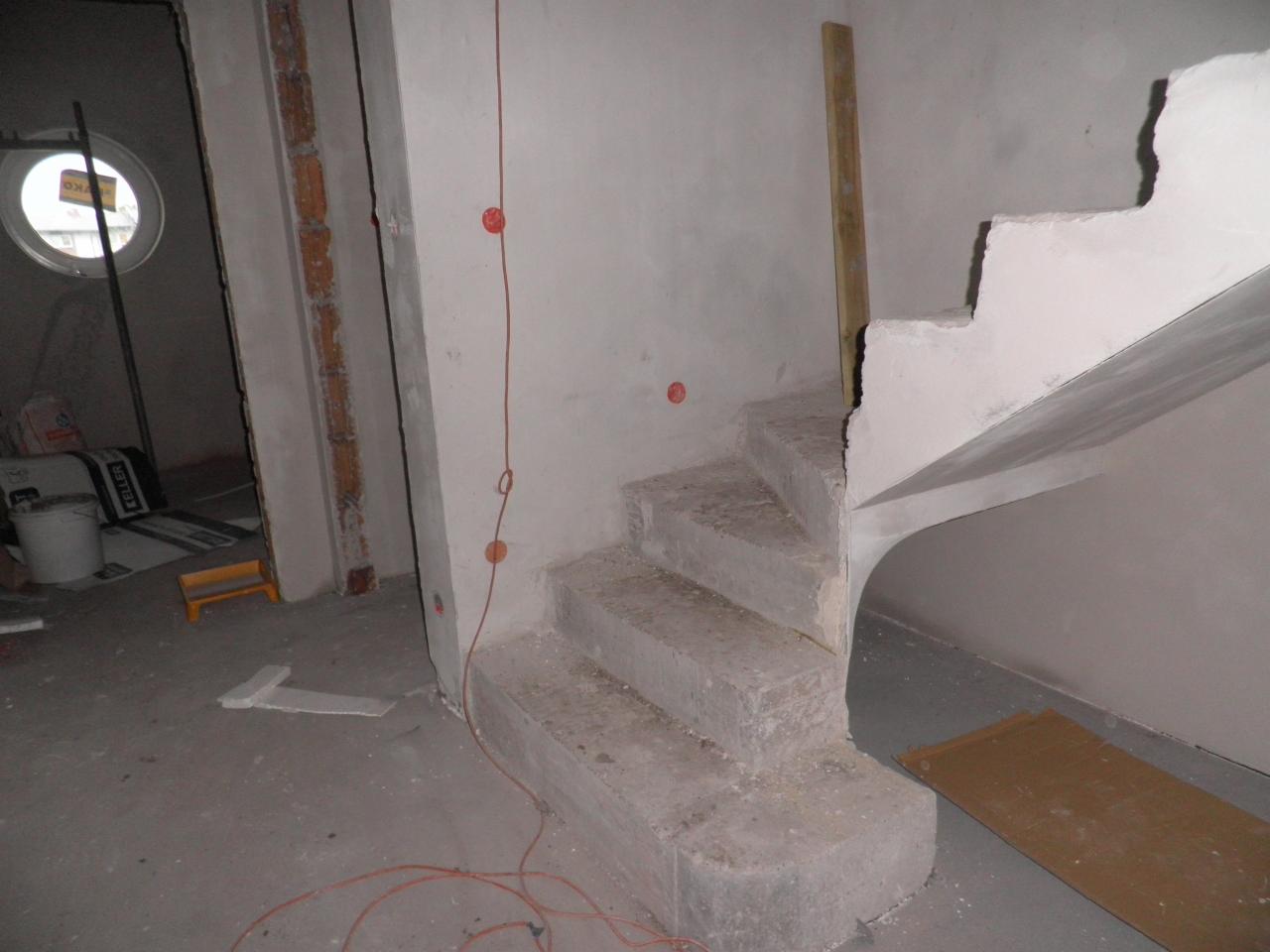 Surowe betonowe schody w trakcie budowy, widoczne niedoskonałości wykończenia, nieotynkowane ściany i wystające kable elektryczne, okrągłe okno w tle.