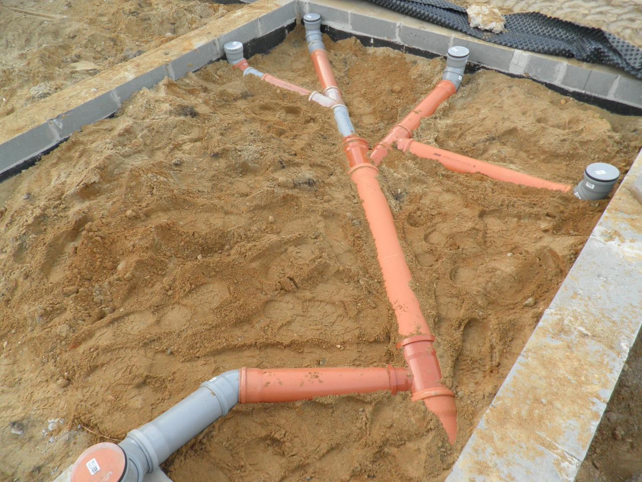 Pomarańczowe i szare rury kanalizacyjne ułożone w ziemi, przygotowane do podłączenia przydomowej oczyszczalni ścieków, widoczne betonowe obrzeża.