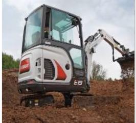 Kompaktowa minikoparka Bobcat E19 w trakcie pracy na placu budowy, z łyżką pełną ziemi, widoczna w tle zieleń.