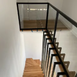 Drewniane schody z balustradą szklaną i czarnymi metalowymi elementami, widok z góry, fragmenty niebieskiej taśmy zabezpieczającej na szkle.