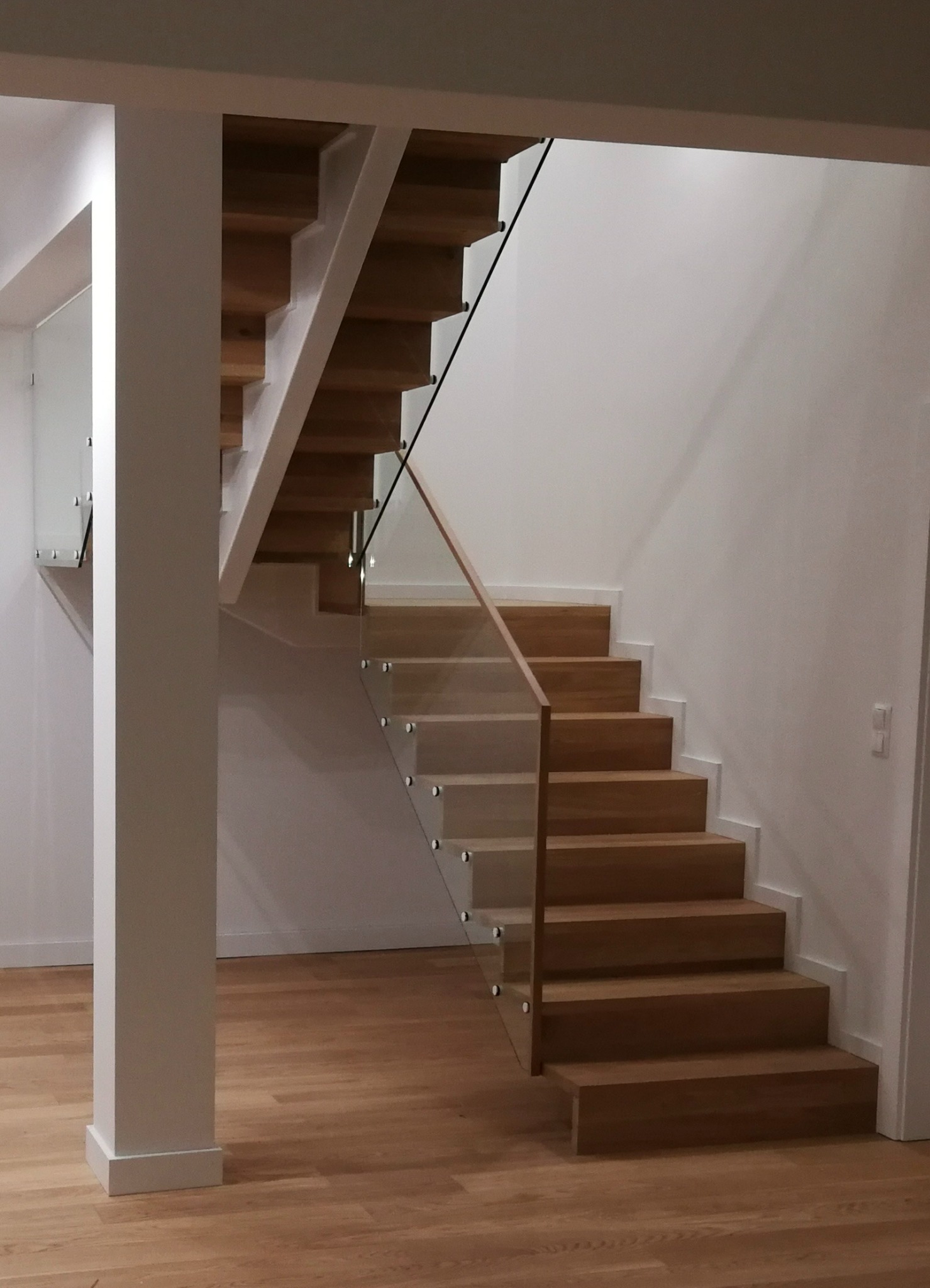 Minimalistyczne schody dywanowe z drewna o jasnym odcieniu, z balustradą ze szkła i drewnianą poręczą, wsparte na białej kolumnie w nowoczesnym wnętrzu.