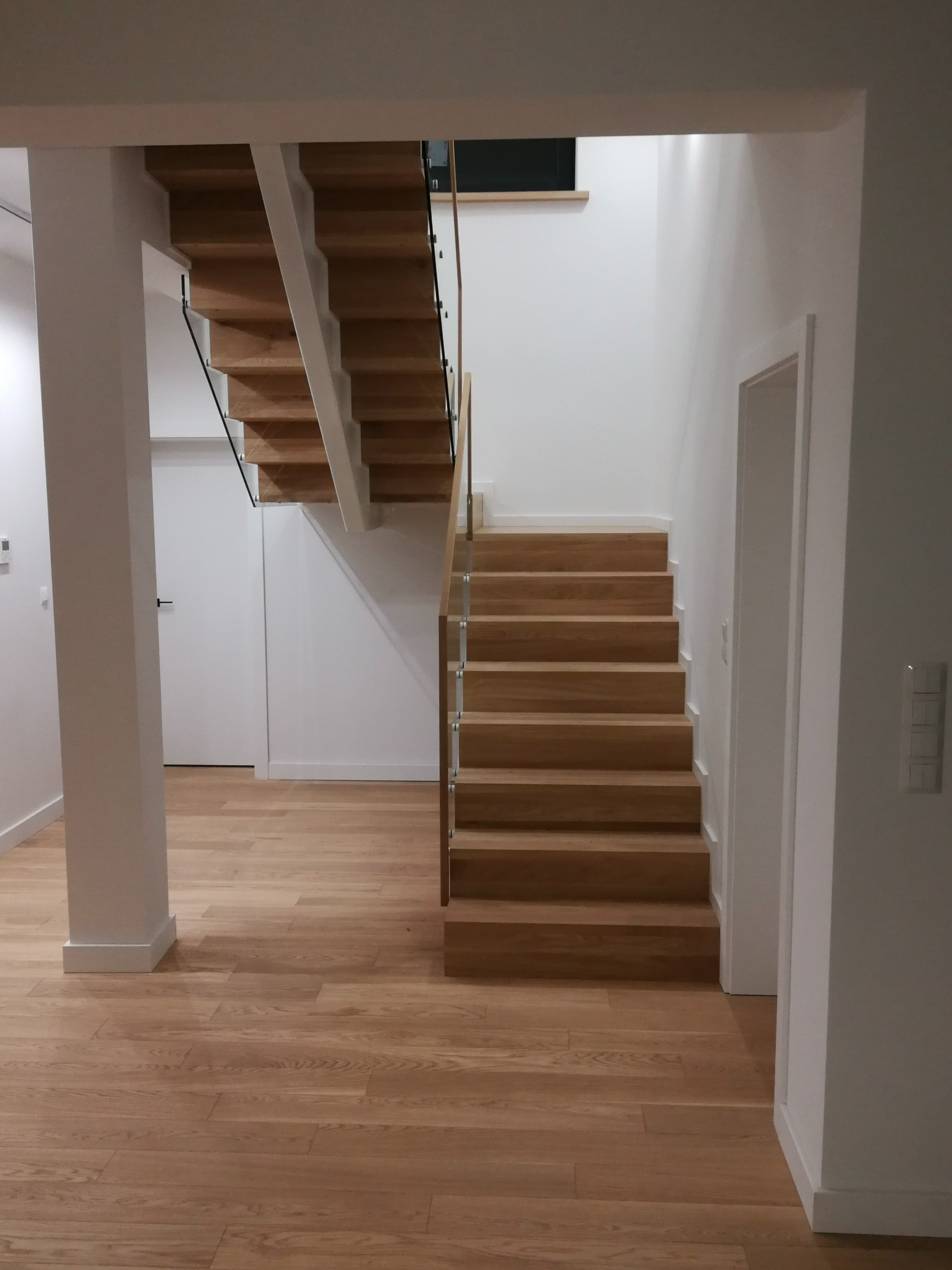 Minimalistyczne schody dywanowe z jasnego drewna z balustradą ze szkła i metalu w nowoczesnym, białym wnętrzu.