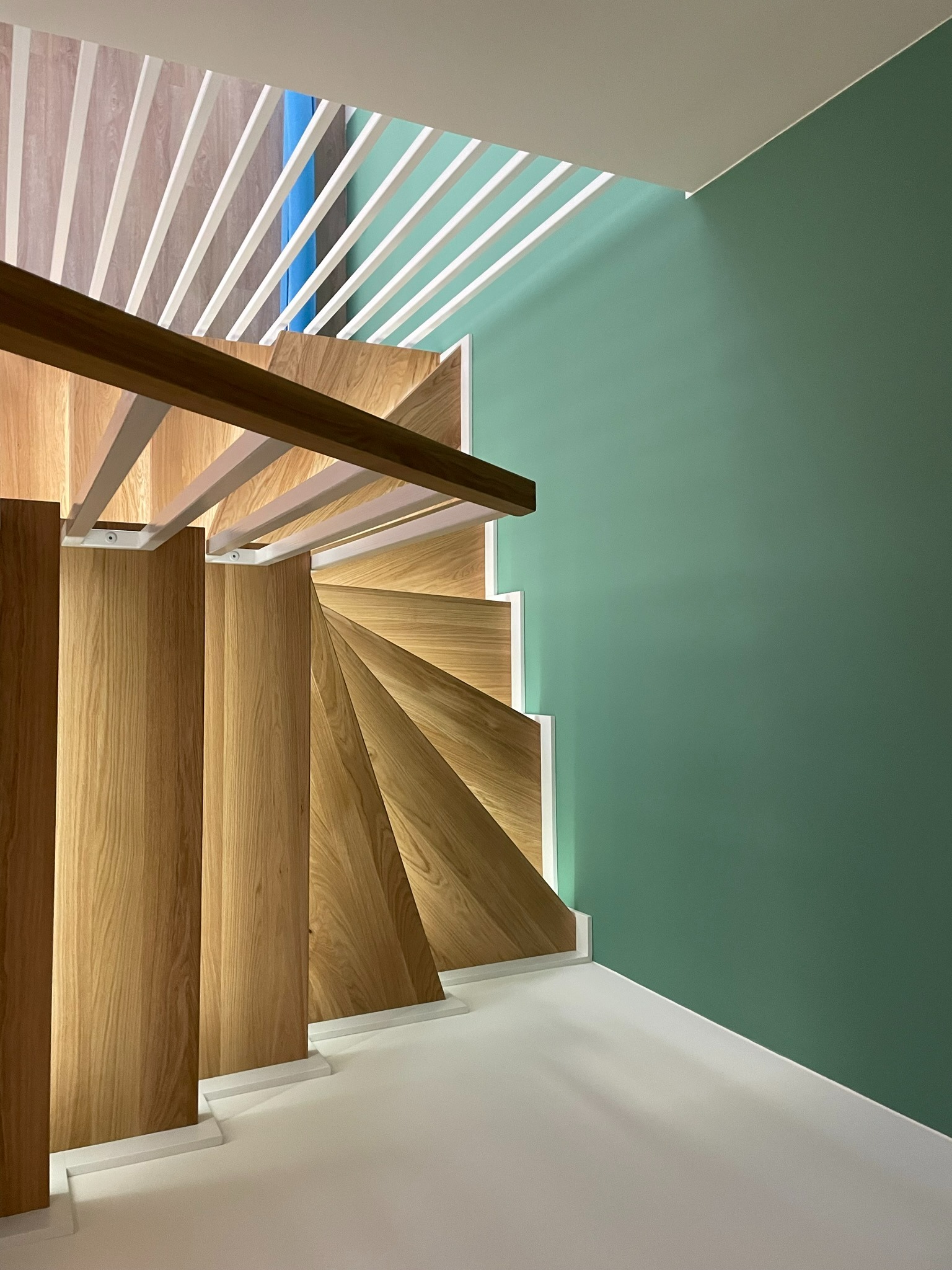 Widok z góry na spiralne schody z jasnego drewna z białymi podstopnicami i balustradą z białych listew, na tle zielonej ściany.
