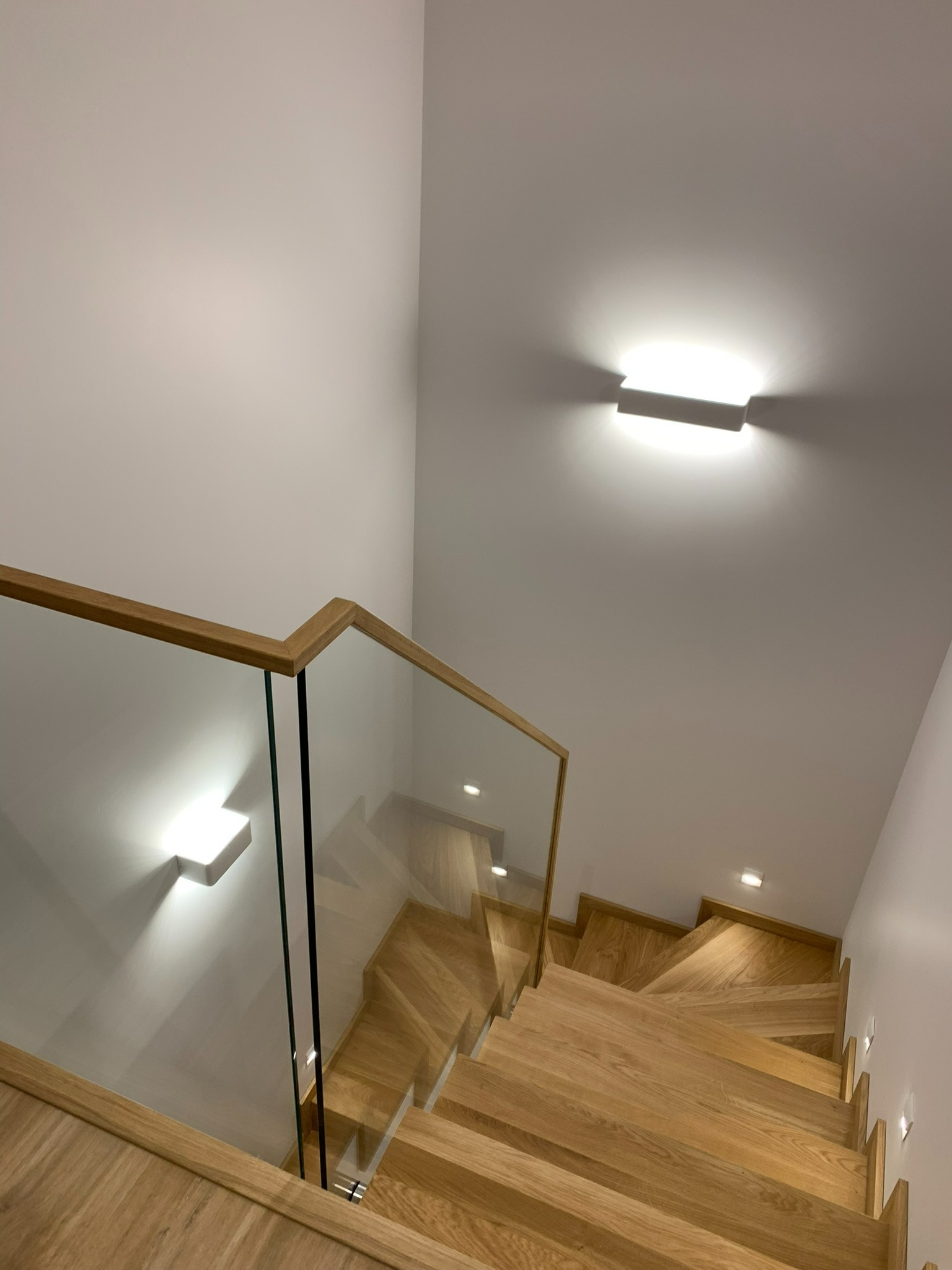Nowoczesne schody z drewna z balustradą ze szkła, oświetlone punktowymi lampami na ścianie, widok z góry.