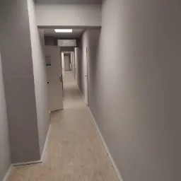 Długi, szary korytarz z beżową podłogą, białymi drzwiami i tabliczką informacyjną 'Kierownik Centrum Sportu', oświetlony kwadratowymi lampami sufitowymi.