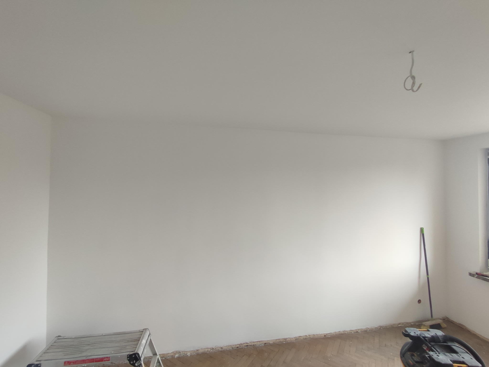 Świeżo pomalowane na biało ściany pokoju z parkietem, widoczna drabina malarska, odkurzacz przemysłowy i hak w suficie.