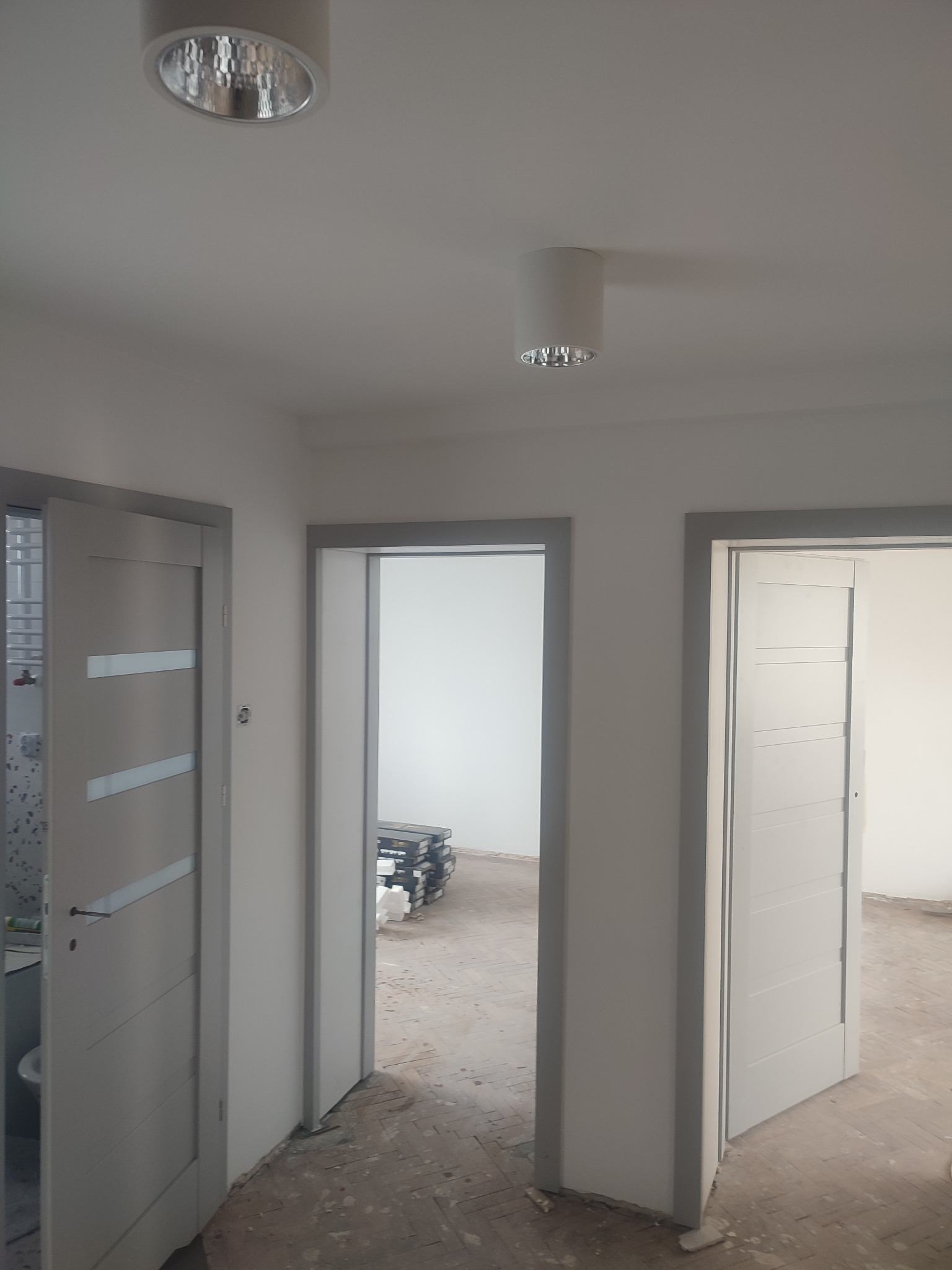Wnętrze mieszkania w trakcie remontu, widoczne trzy otwarte drzwi z szarymi ościeżnicami, białe ściany i sufit z zamontowanymi lampami punktowymi, podłoga częściowo zakryta folią i resztkami...