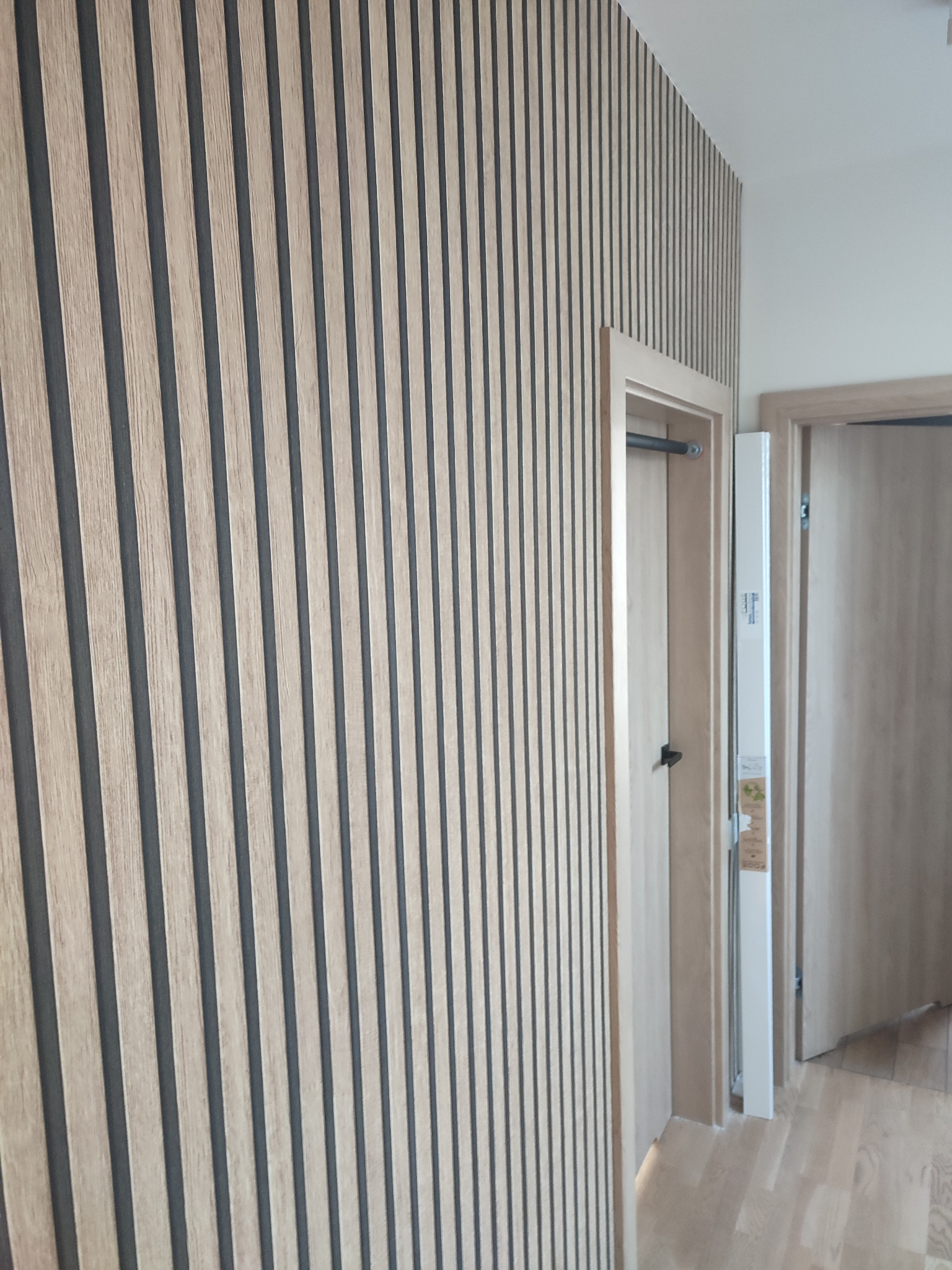 Ściana wykończona panelami ściennymi w odcieniach drewna i grafitu, widoczne drzwi z ościeżnicą w jasnym drewnie oraz fragment podłogi z paneli drewnianych.