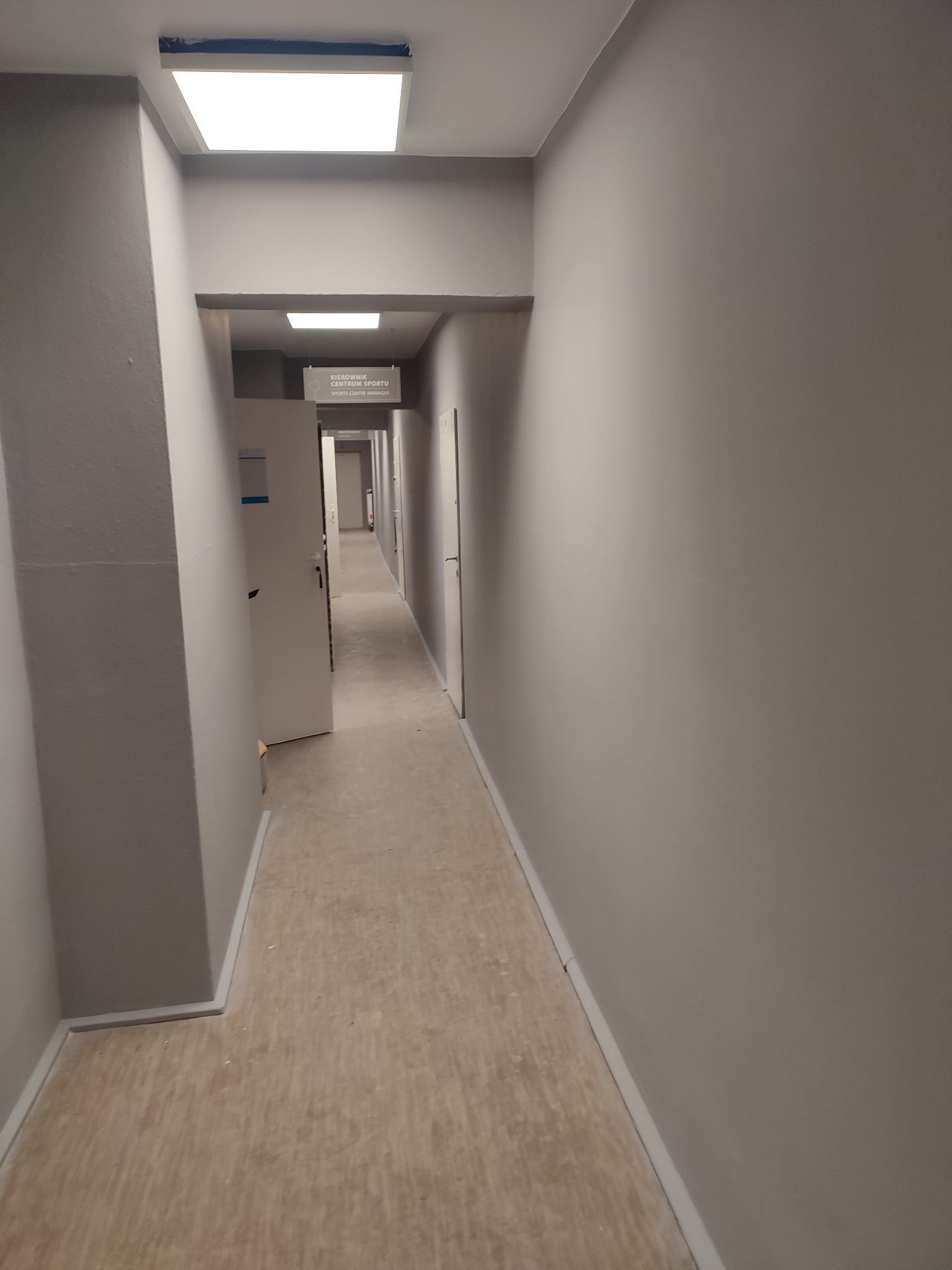 Długi, szary korytarz z beżową podłogą, białymi drzwiami i tabliczką informacyjną 'Kierownik Centrum Sportu', oświetlony kwadratowymi lampami sufitowymi.