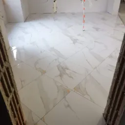 Białe płytki podłogowe imitujące marmur, ułożone w kwadraty, widoczne z perspektywy przejścia w stanie surowym.