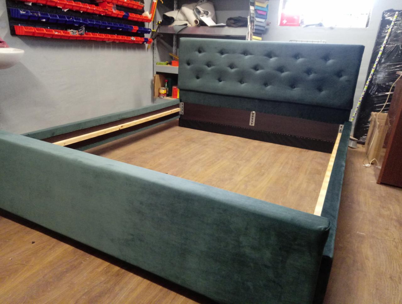 Rama łóżka z tapicerowanym zagłówkiem w odcieniach zieleni, widoczne drewniane elementy konstrukcyjne, tło warsztatowe z narzędziami i akcesoriami.