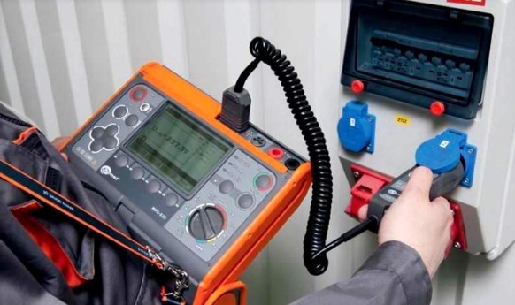 Elektryk w trakcie pracy, używa miernika MFT-535 do sprawdzenia gniazdka elektrycznego w skrzynce rozdzielczej. Widoczny wyświetlacz miernika.