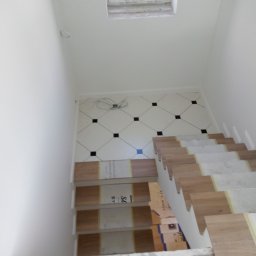 WAW-GLAZ - Białe płytki podłogowe z czarnymi akcentami i drewnopodobne stopnie schodów w trakcie montażu, widok z góry, okno dachowe w tle.