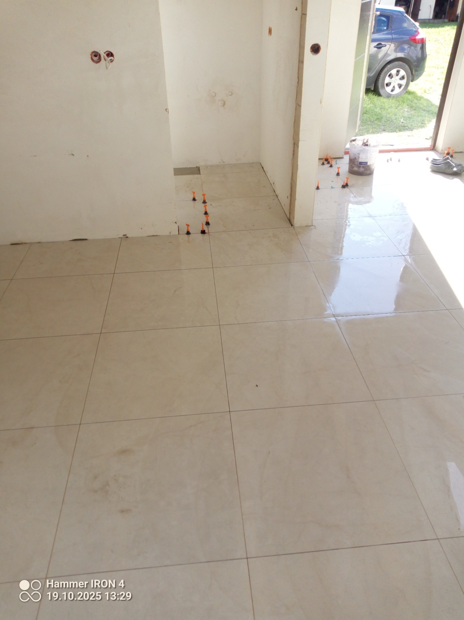 Jasne, kwadratowe płytki podłogowe w trakcie układania, z pomarańczowymi krzyżykami dystansowymi. Widoczne ślady po fugowaniu i fragment ściany z instalacją elektryczną.