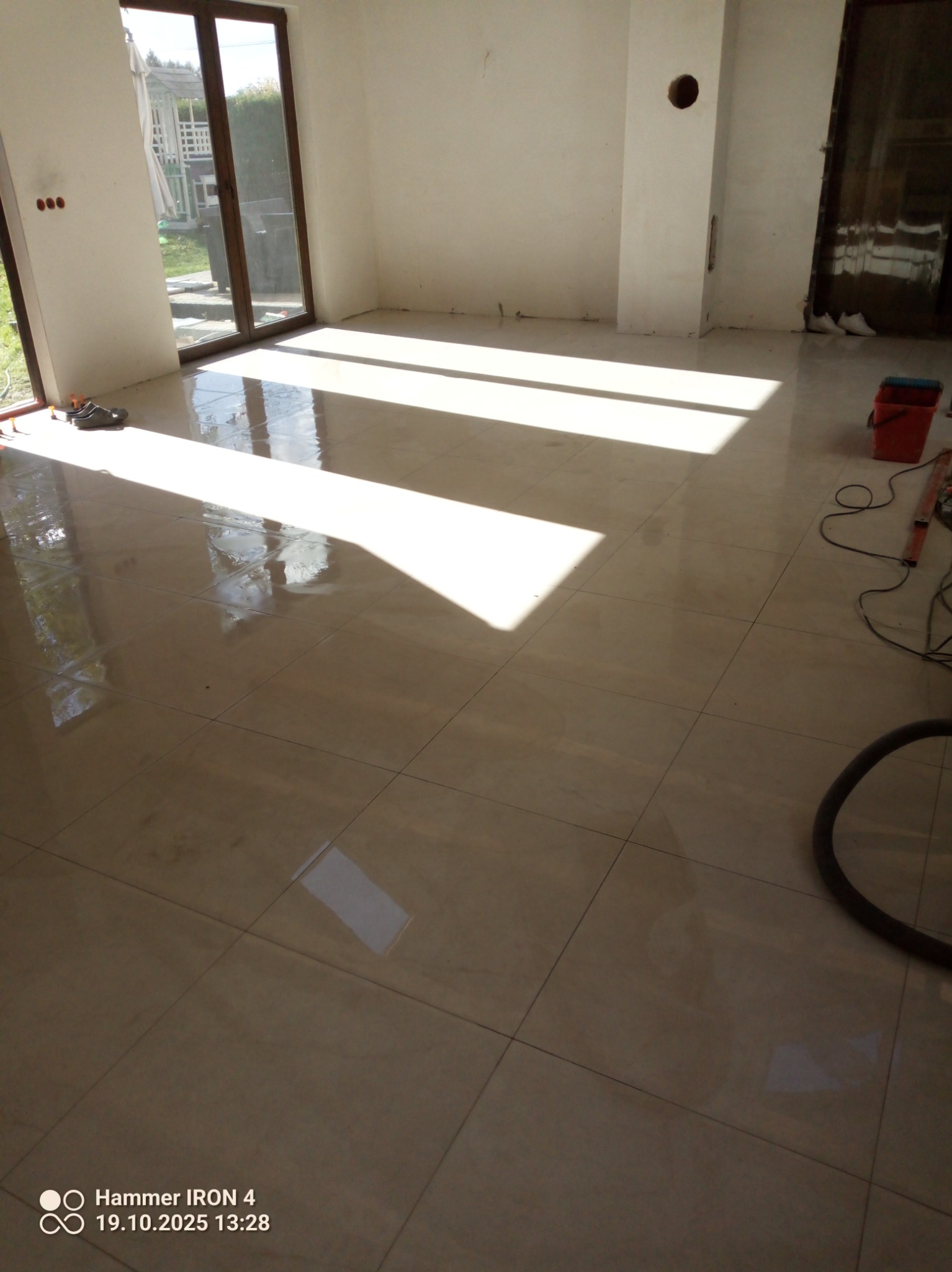 Błyszcząca, beżowa podłoga z płytek w nowym wnętrzu. Widoczne narzędzia pracy, wiadro i węże odkurzacza. Promienie słoneczne wpadają przez okno.
