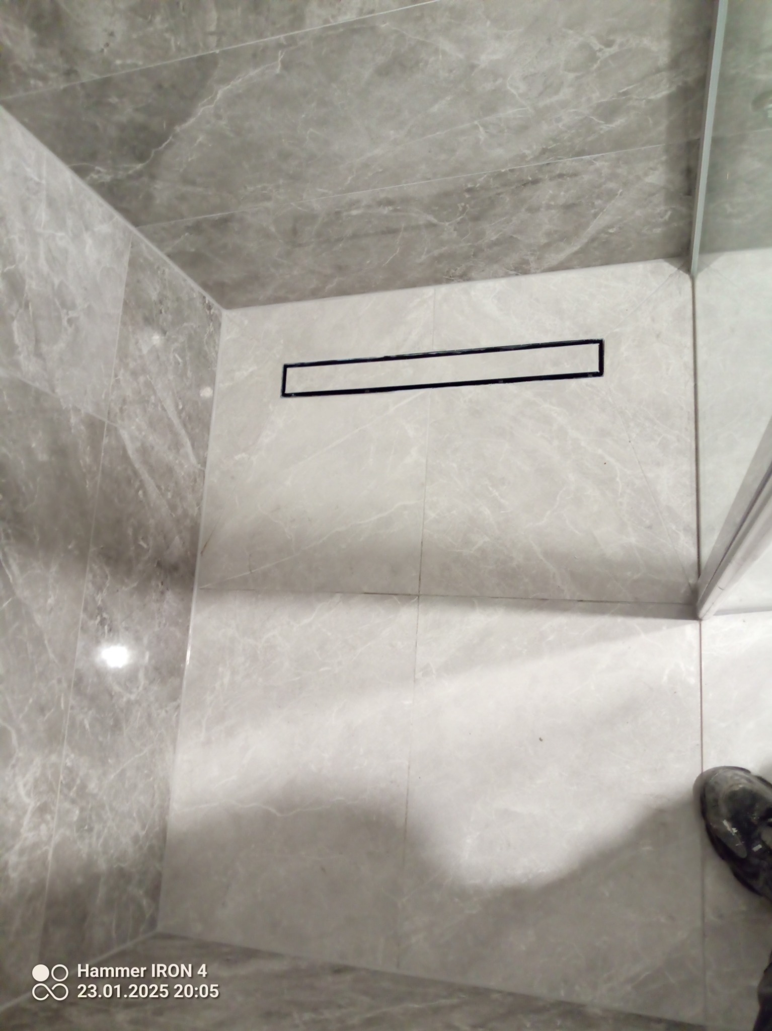 Wykończona kabina prysznicowa z odpływem liniowym i szarymi płytkami imitującymi marmur. Widoczny fragment buta w rogu kadru.