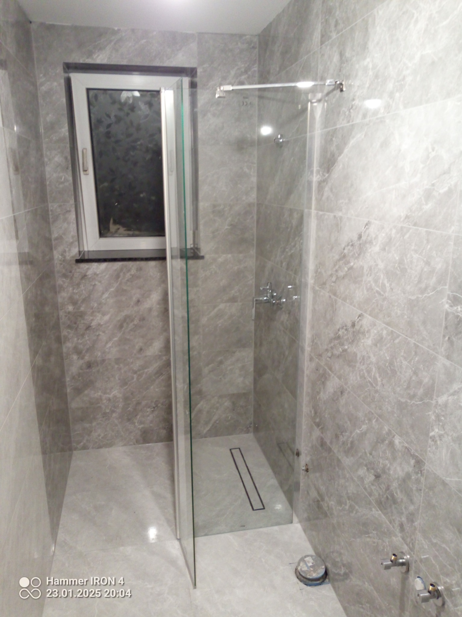 Szara łazienka z kabiną prysznicową typu walk-in, wykończona marmurowymi płytkami. Widoczny odpływ liniowy i okno z widokiem na zieleń. Minimalistyczny design.