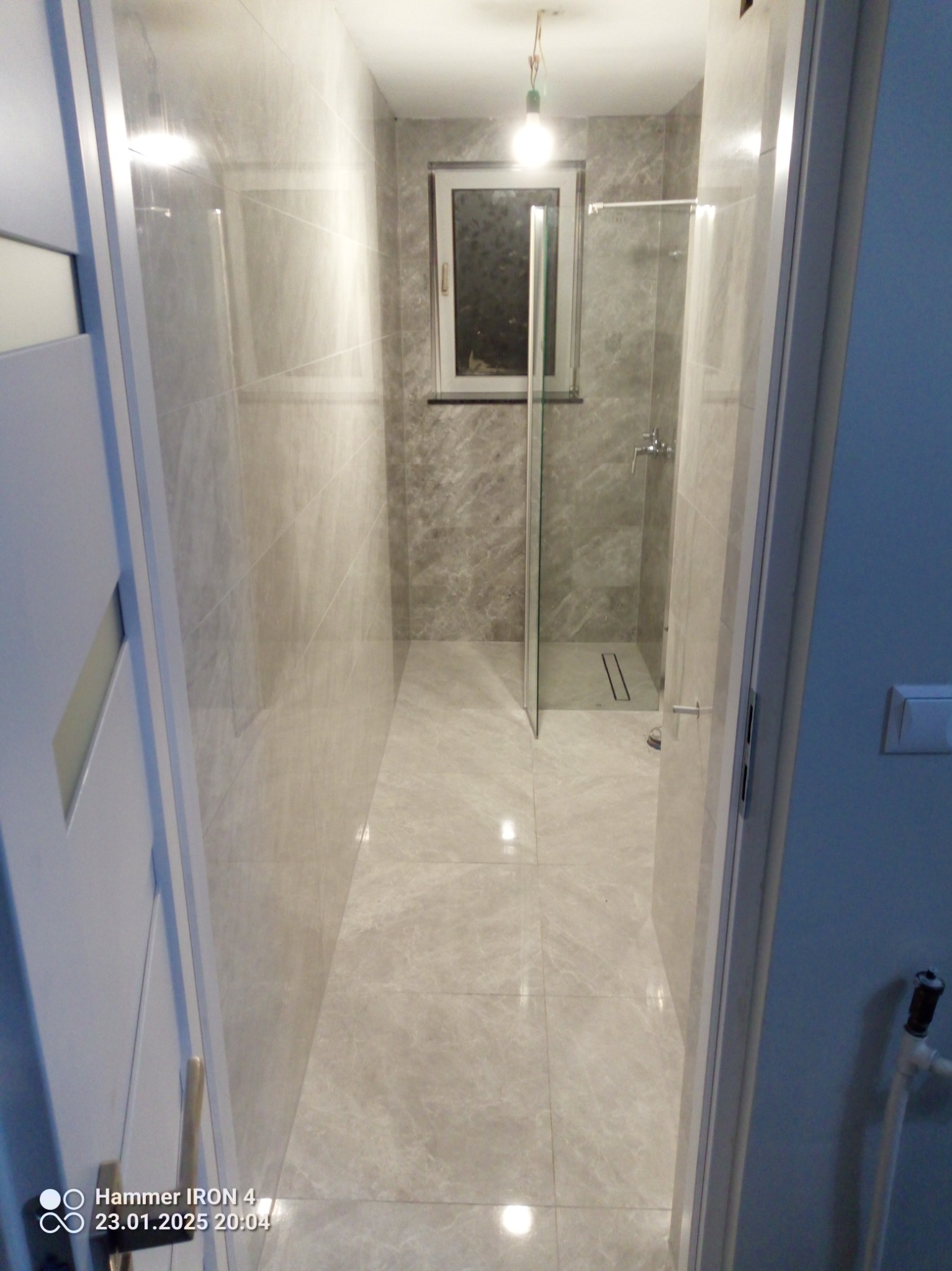 Wykończona łazienka z kabiną prysznicową typu walk-in, wyłożona jasnymi płytkami imitującymi marmur. Widoczne okno i oświetlenie sufitowe. Minimalistyczny design.