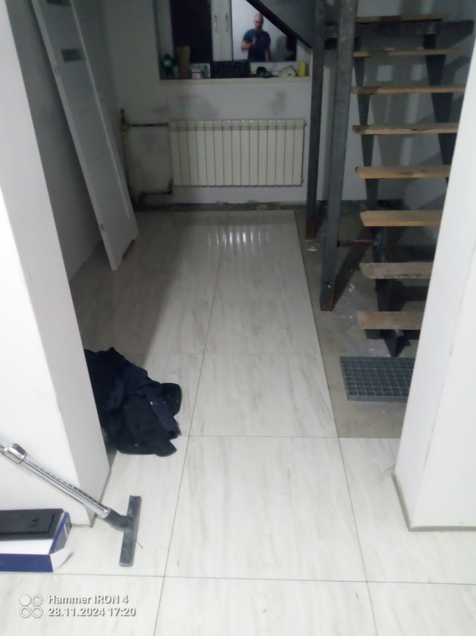 Wykończona podłoga z płytek w jasnym odcieniu, widok na schody i okno z odbiciem osoby. Na podłodze leżą narzędzia i ubrania, co sugeruje trwające prace wykończeniowe.