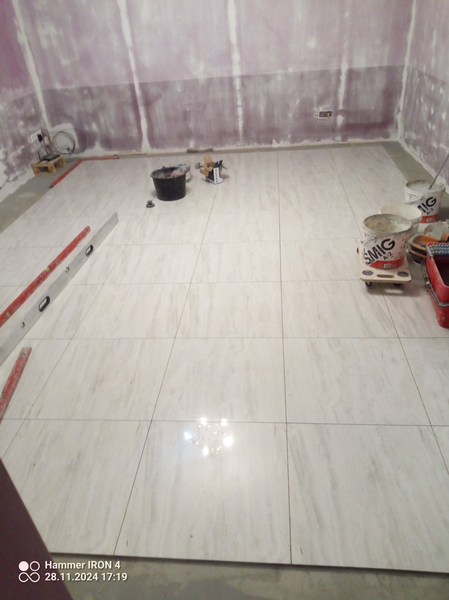 Ułożone jasne płytki podłogowe w trakcie remontu łazienki. Widoczne narzędzia glazurnicze, poziomica i wiaderka z zaprawą. Ściany przygotowane do dalszych prac wykończeniowych.