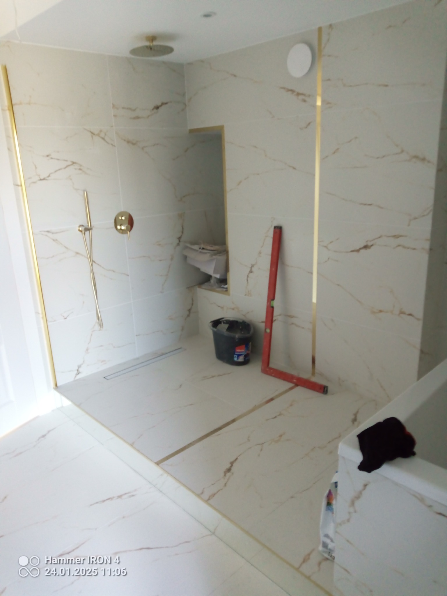 Wykończona łazienka z beżowymi płytkami imitującymi marmur, złotymi listwami i deszczownicą. Widoczny poziomica i wiadro, co sugeruje trwające prace wykończeniowe.