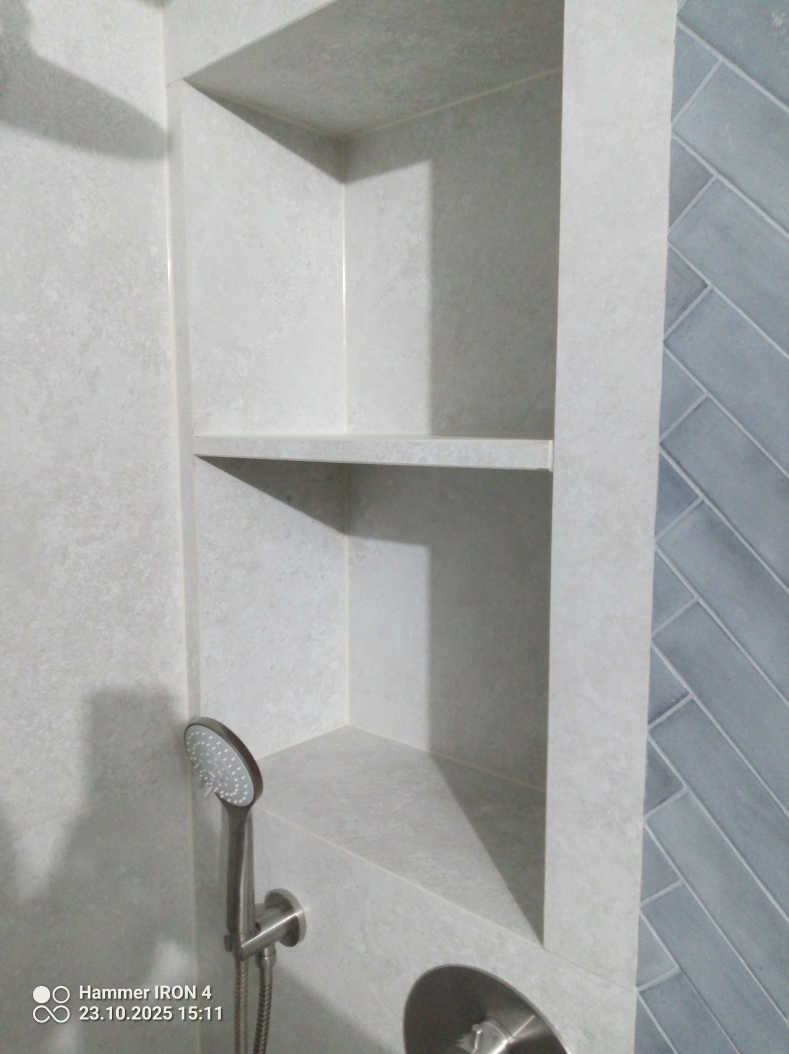 Wnęka z półkami wyłożona jasnymi płytkami w łazience, obok ściana z niebieską glazurą ułożoną w jodełkę. Widoczna bateria i słuchawka prysznicowa.