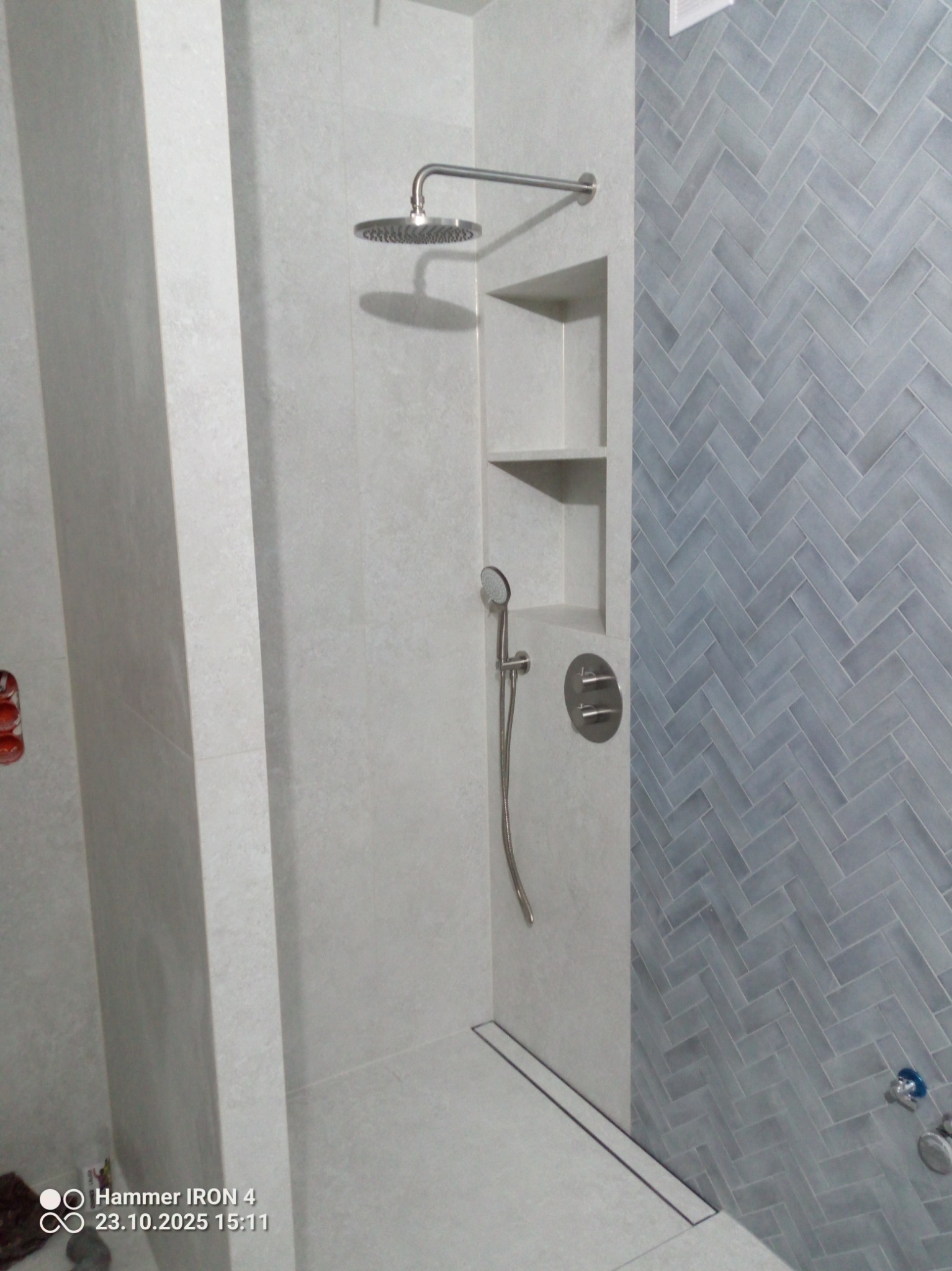 Wykończona kabina prysznicowa z szarymi płytkami ułożonymi w jodełkę, wnęką z półkami i odpływem liniowym. Nowoczesny design, minimalistyczny styl.