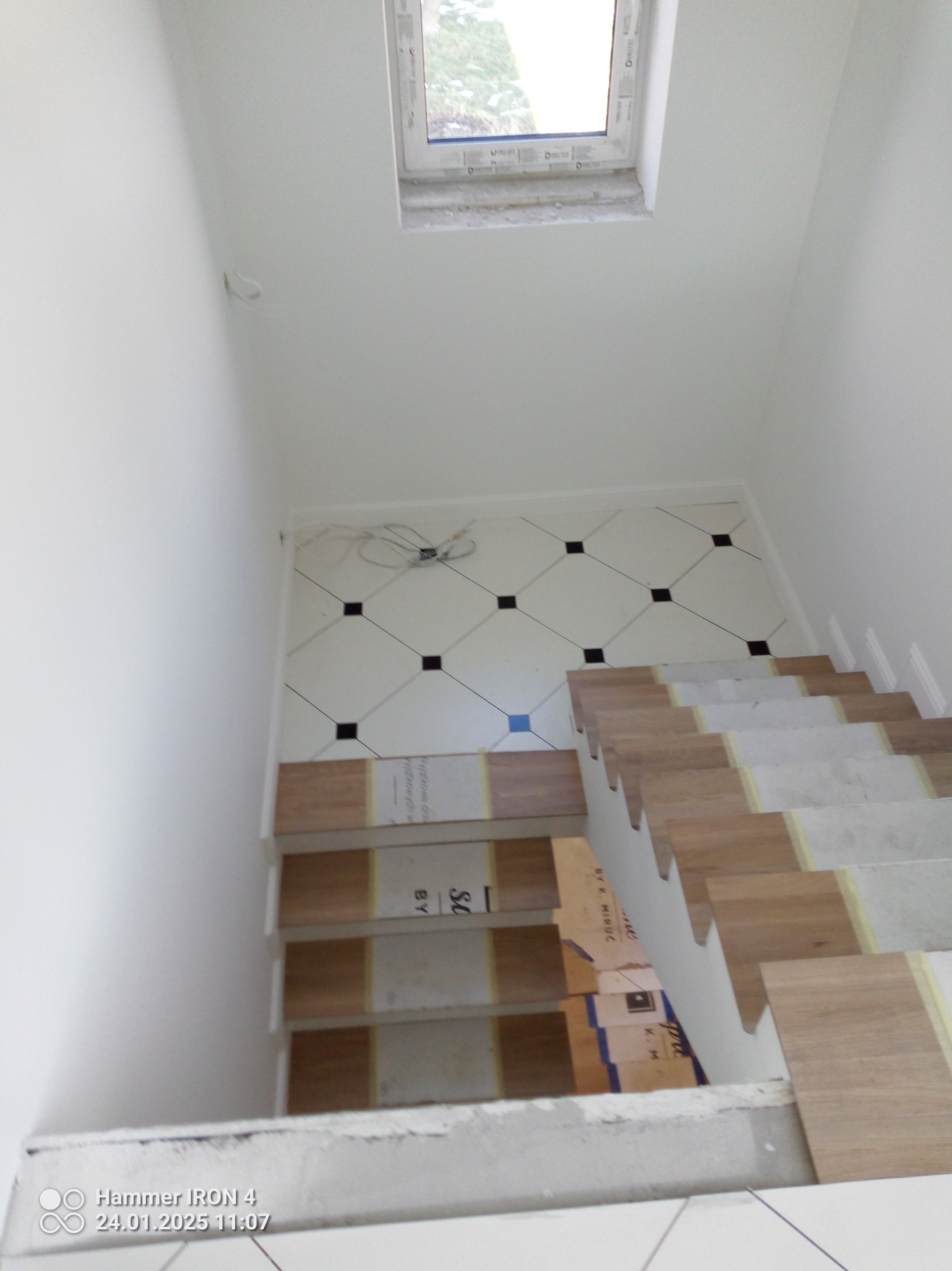 Białe płytki podłogowe z czarnymi akcentami i drewnopodobne stopnie schodów w trakcie montażu, widok z góry, okno dachowe w tle.