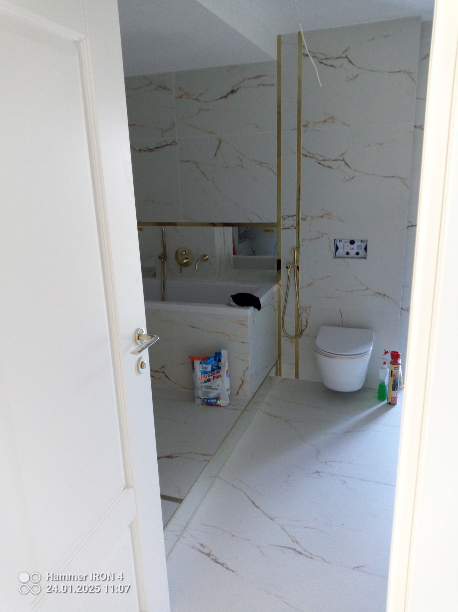 Wykończona łazienka z wanną, toaletą i złotymi akcentami, widoczna przez uchylone białe drzwi. Na podłodze i ścianach płytki imitujące marmur.