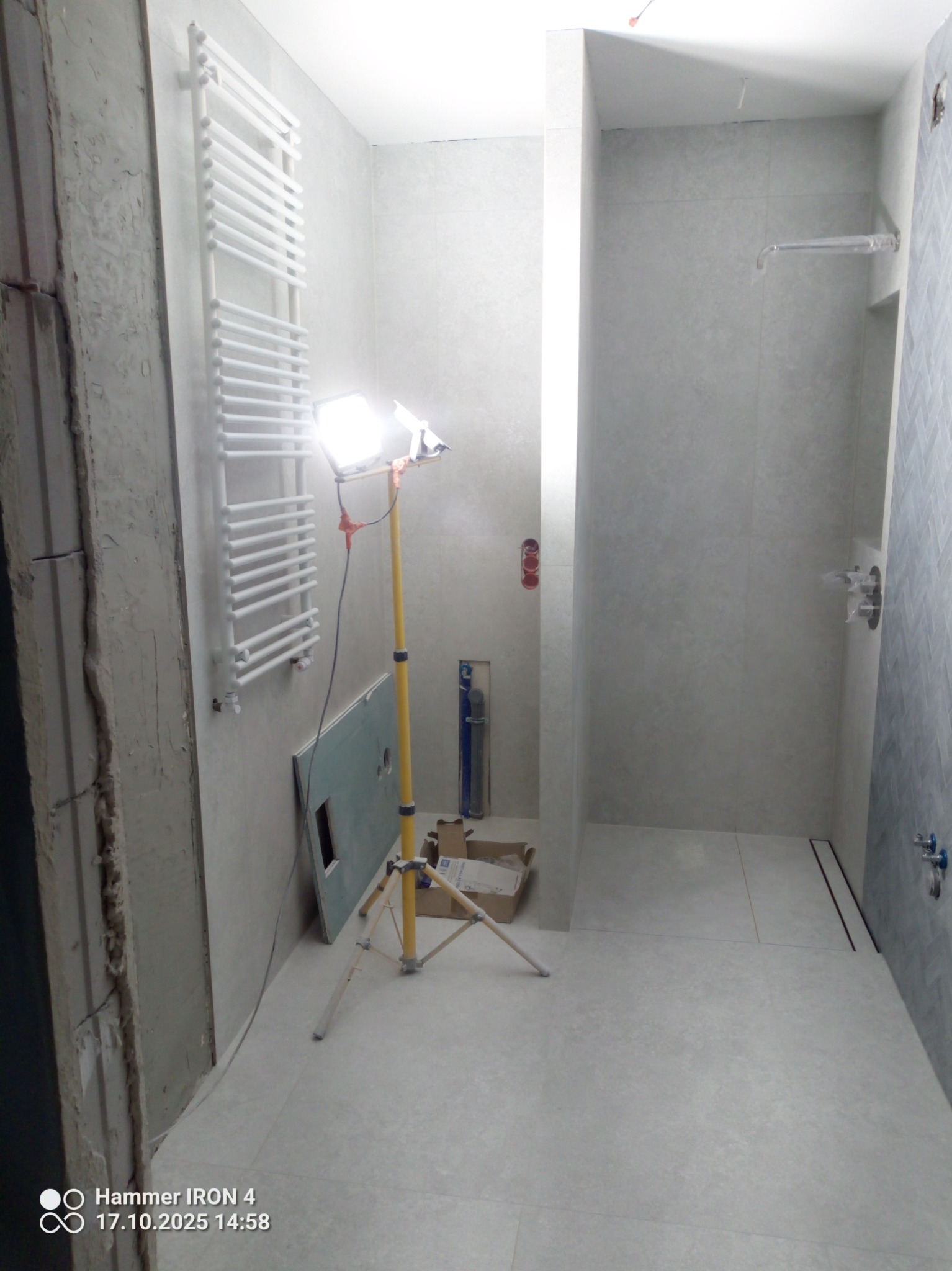 Wnętrze łazienki w trakcie prac wykończeniowych: ułożone płytki, zamontowany grzejnik drabinkowy, widoczna lampa budowlana i materiały instalacyjne.