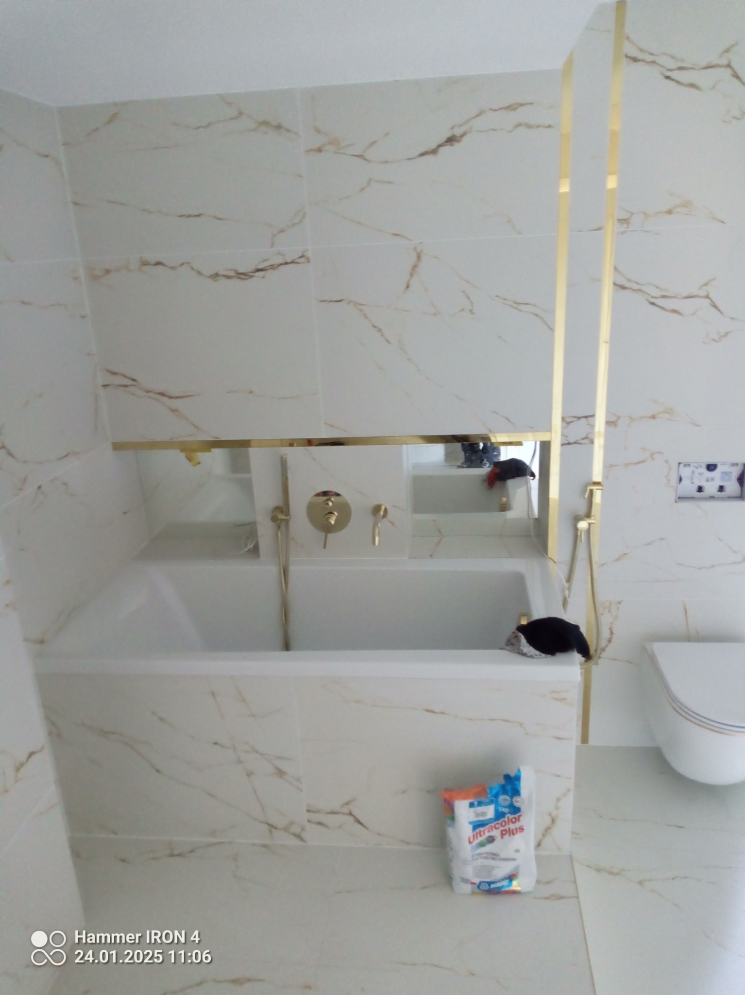 Wykończona łazienka z wanną, złotymi akcentami i marmurowymi płytkami. Widoczny fragment toalety i opakowanie fugi. Minimalistyczny design z dbałością o detale.