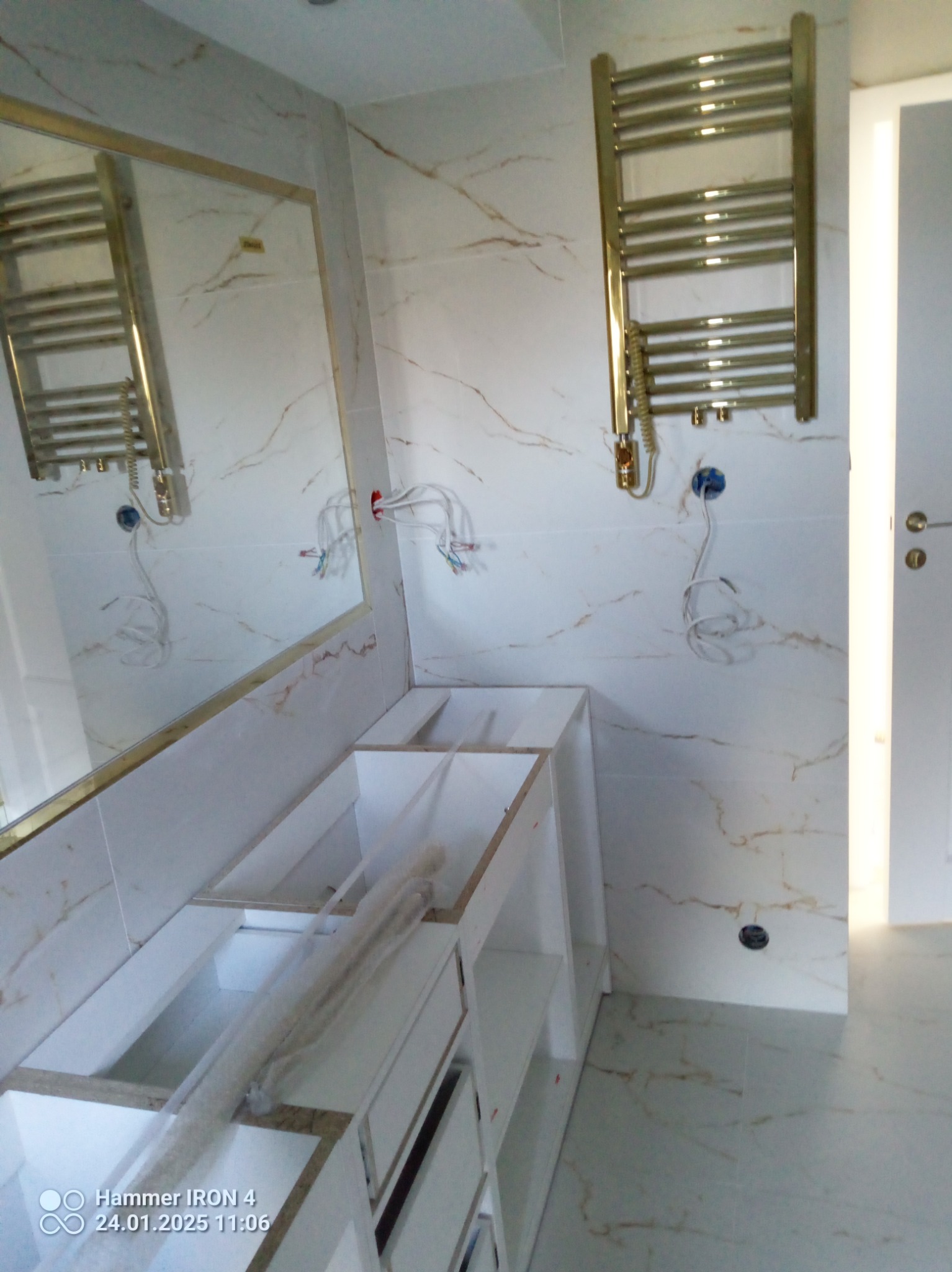 Łazienka w trakcie wykończenia: białe marmurowe płytki, złoty grzejnik drabinkowy, lustro w złotej ramie i biała szafka pod umywalkę w trakcie montażu.