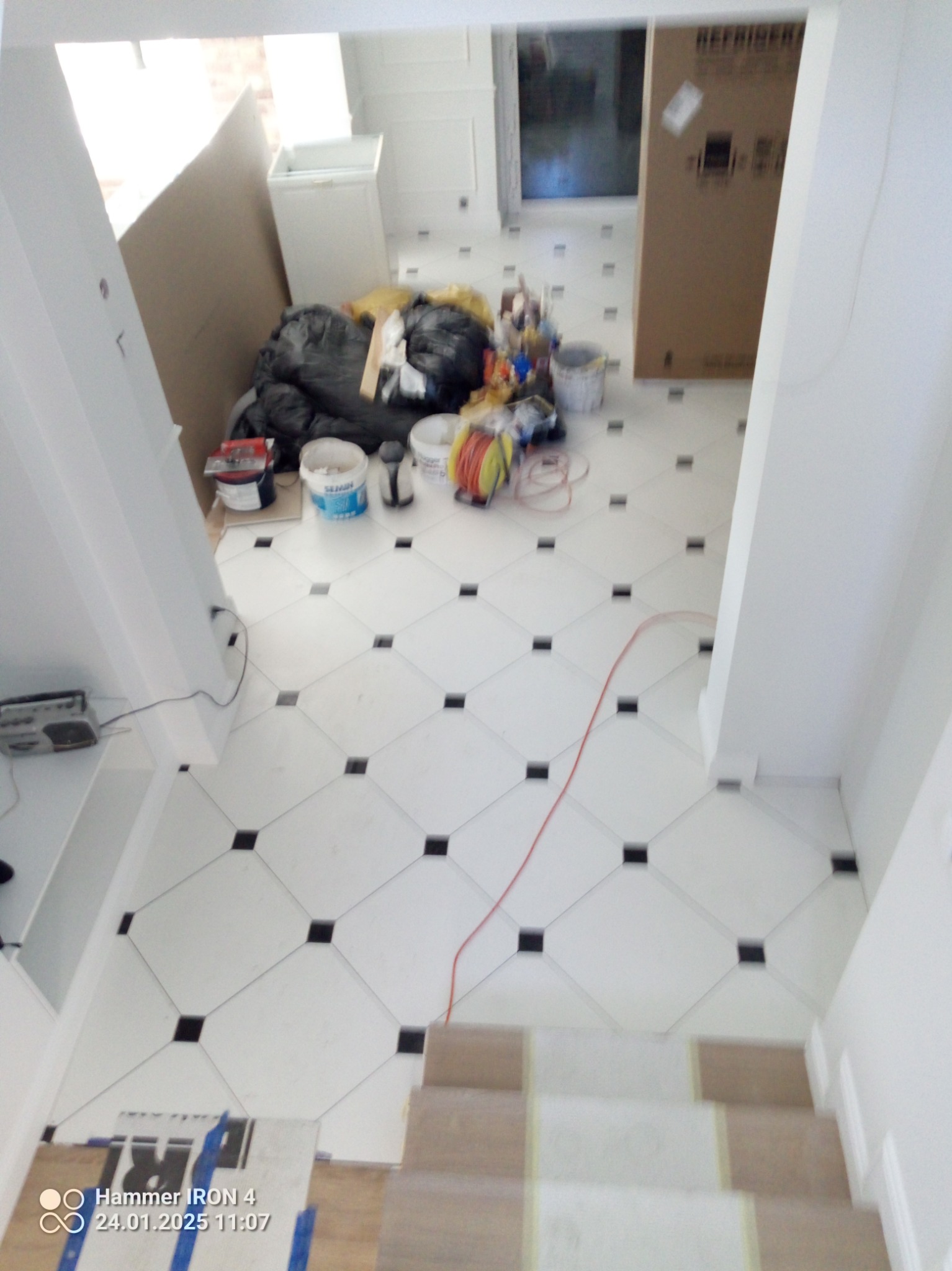 Biała podłoga z kwadratowych płytek z czarnymi akcentami, widok z góry na schody, w tle narzędzia i materiały budowlane. Ukończony fragment podłogi.